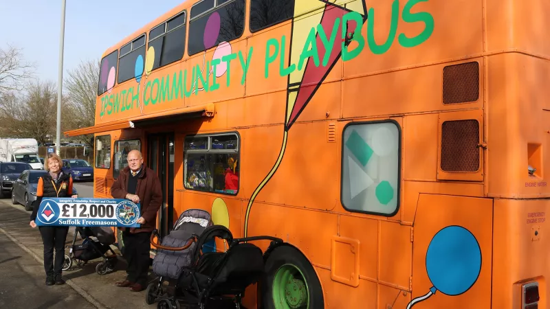 two people stand outside a bus holding a banner that says '£12,000 Suffolk Freemasons'. The Bus is a bright orange double decker bus with Ipswitch Community Playbus written in green writing. There are balloons and a kit painted onto the side.