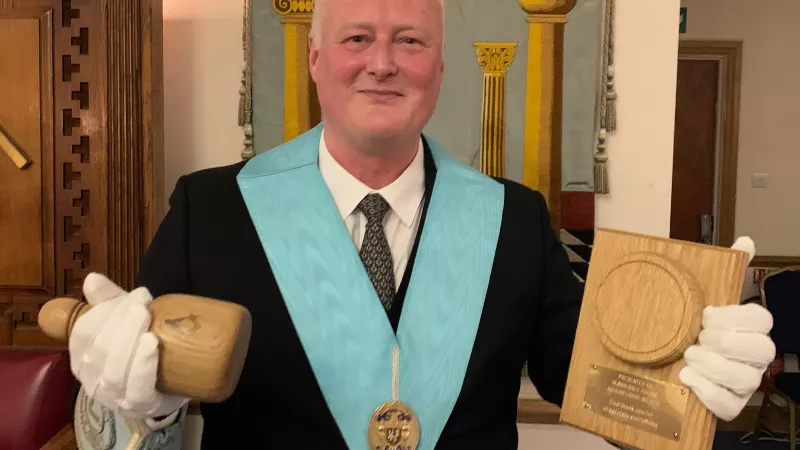A middle aged man in freemasonry regalia is holding a wooden gavel with a plaque and inscription.