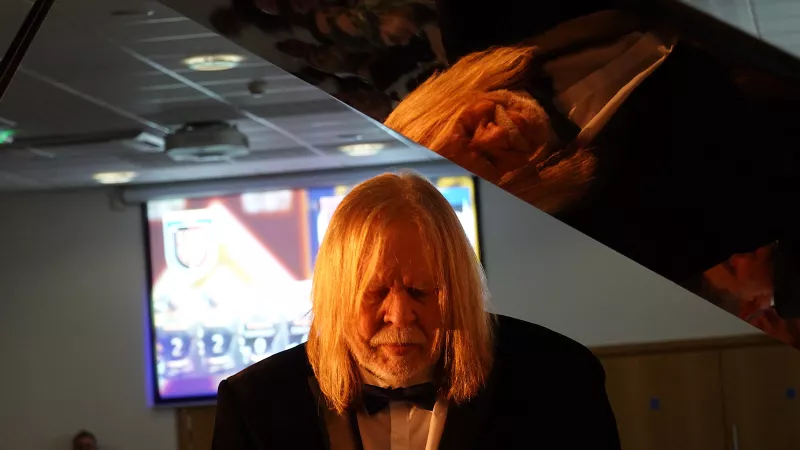 An older white man with long light coloured heair and beard, sits playing a grand piano wearing black tie. His reflection can be seen in the lid of the piano.