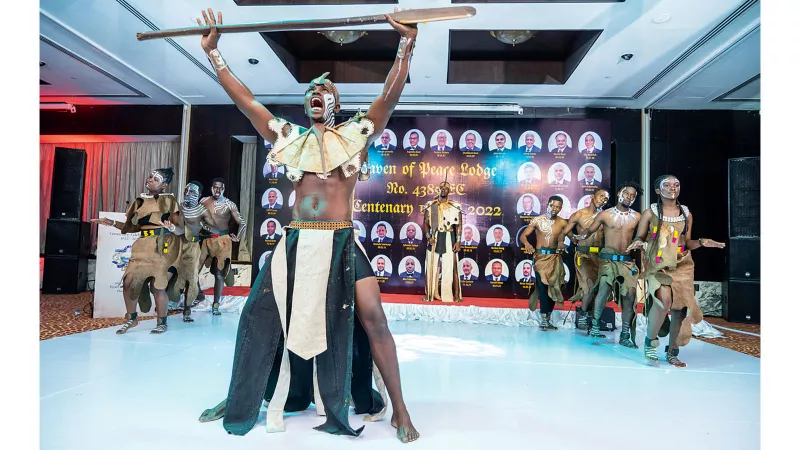 Dancers displaying a traditional dance of cultural significance. A black man with white markings on his face stands in a powerful posture, holding a wooden paddle above his head.