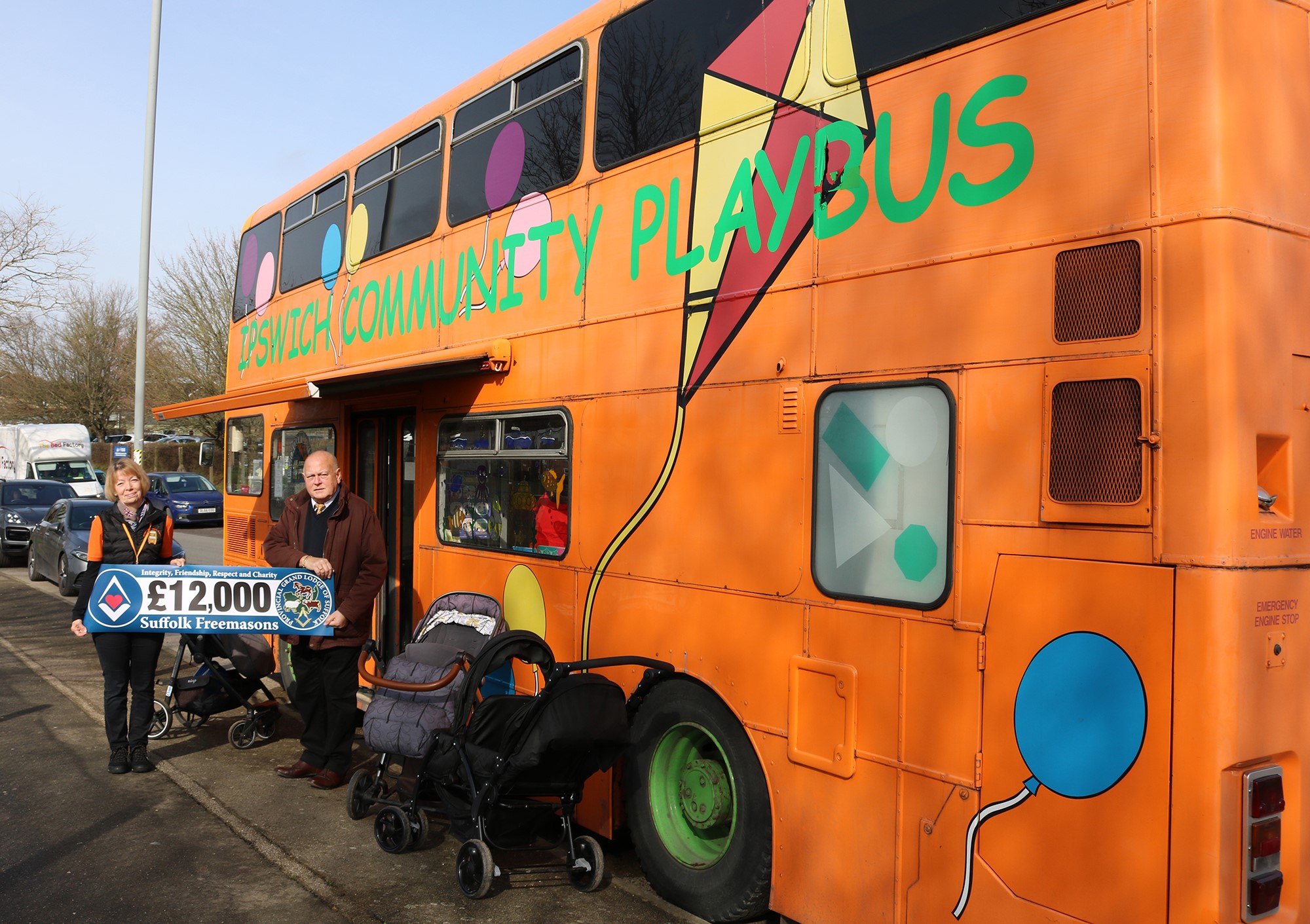two people stand outside a bus holding a banner that says '£12,000 Suffolk Freemasons'. The Bus is a bright orange double decker bus with Ipswitch Community Playbus written in green writing. There are balloons and a kit painted onto the side.