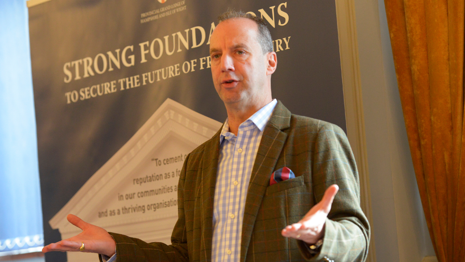 John Whitaker is photographed while giving a speech, he is wearing a green tweed jacket on top of a blue gingham shirt.