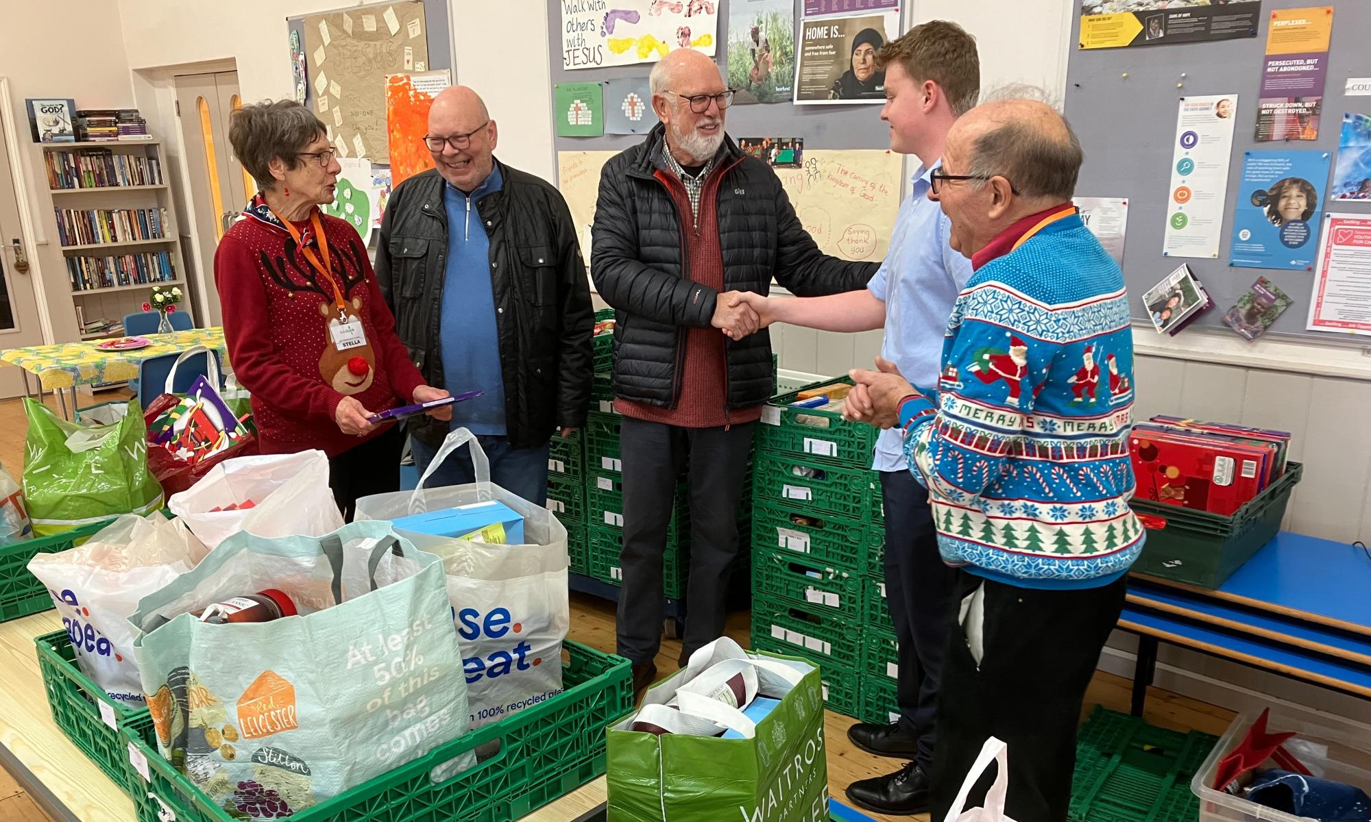 A woman and 5 men standing behind a table which has crates and banks of food items that have been donated to the food bank