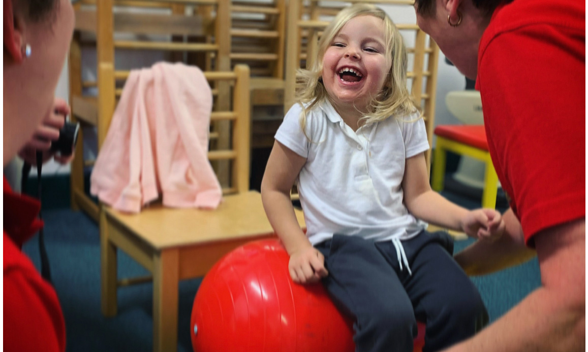 Grace, a young blonde girl, sits on a red yoga ball, she is laughing.