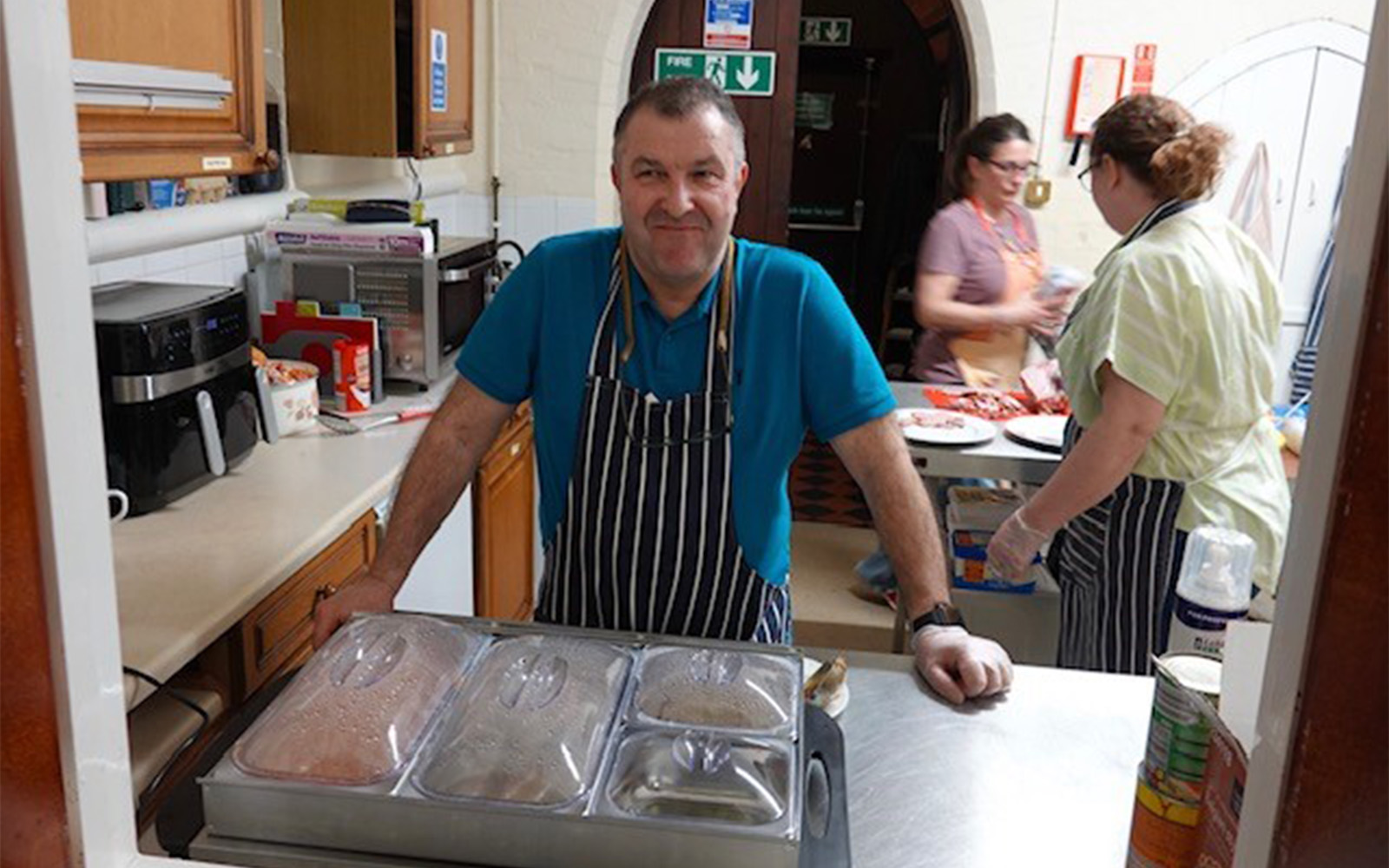 Man stood in kitchen area with covered catering dishes of food in front of him.
