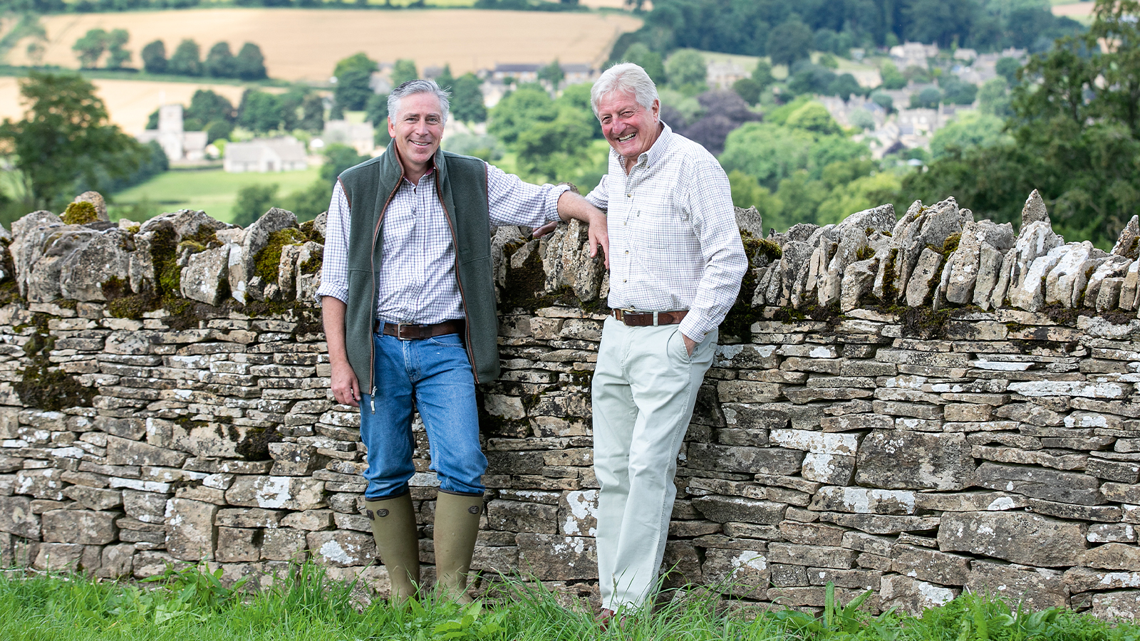 Tony and Tony2 stand in front of an old stone wall, they are relaxed and smiling at the camera.