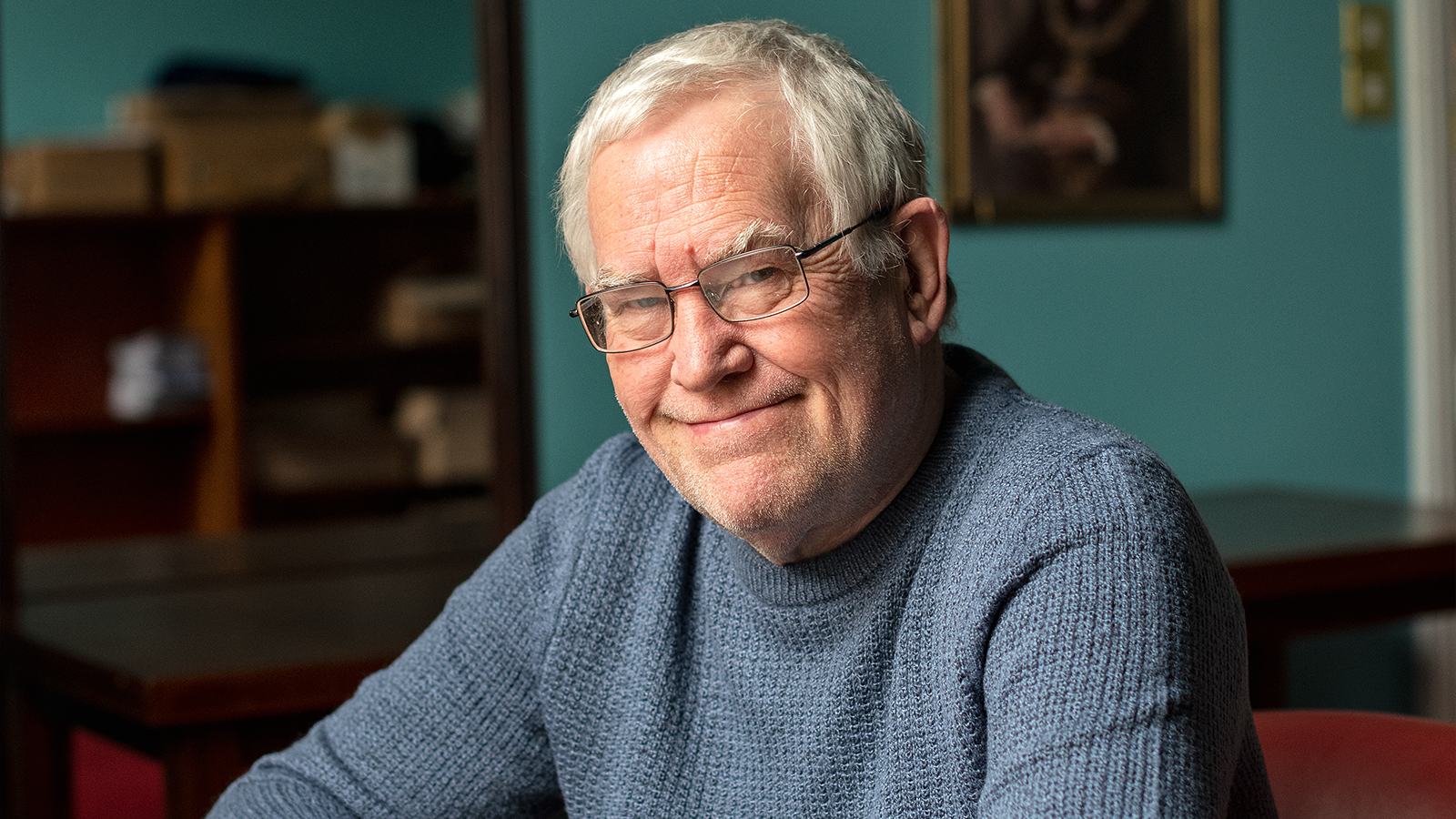 Gary is pictured with a blue knitted jumper, he is wearing rectangular glasses with a thing metal frame and is looking at the camera and smiling.
