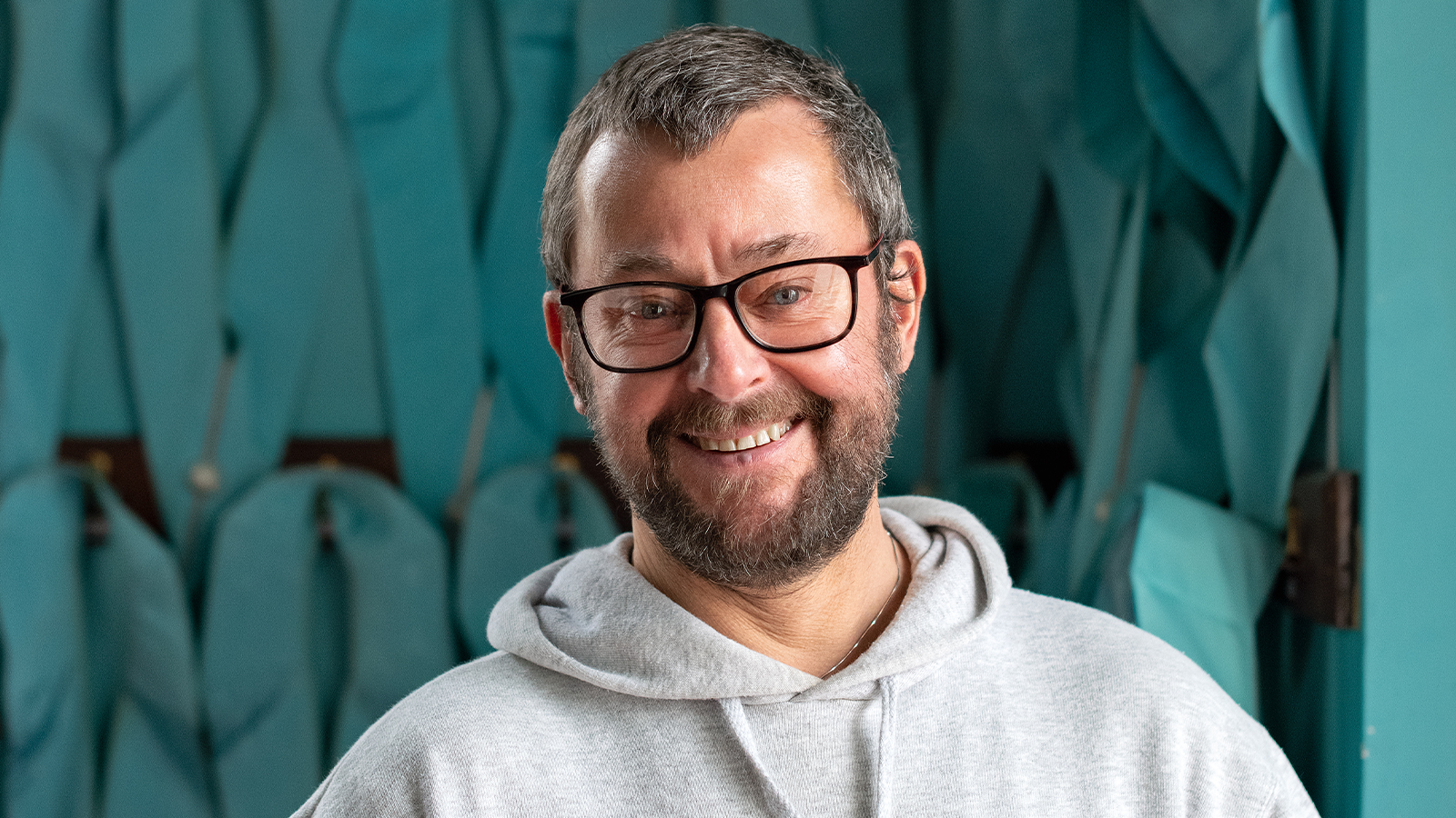 Steven is a bearded man, wearing a light grey hoodie and black, thick rimmed glasses. He is smiling at the camera.