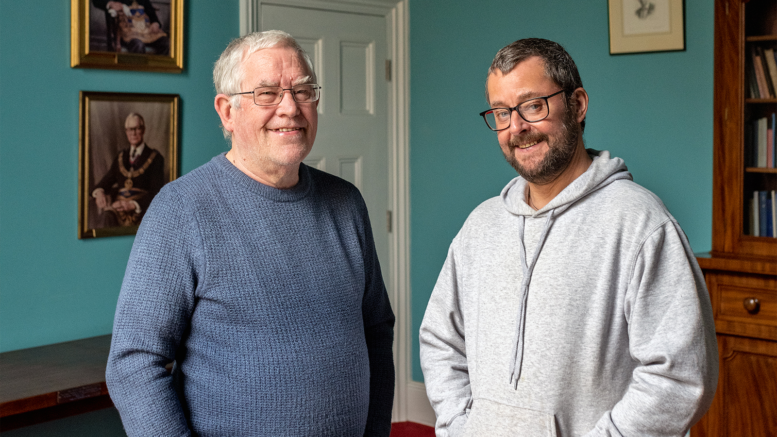 Gary and Steven Standing in a room with bright blue walls, they both wear glasses and are smiling and the camera.