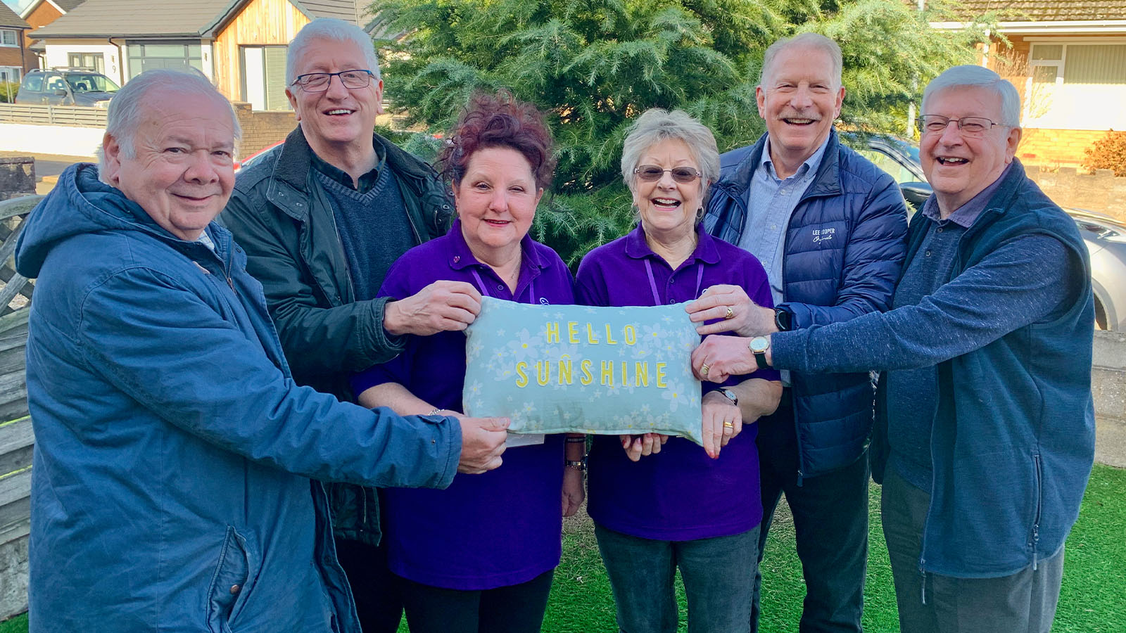 Six people standing in a group holding a blue rectangular cushion with the words 'HELLO SUNSHINE' written on it in gold. They are all smiling at the camera.