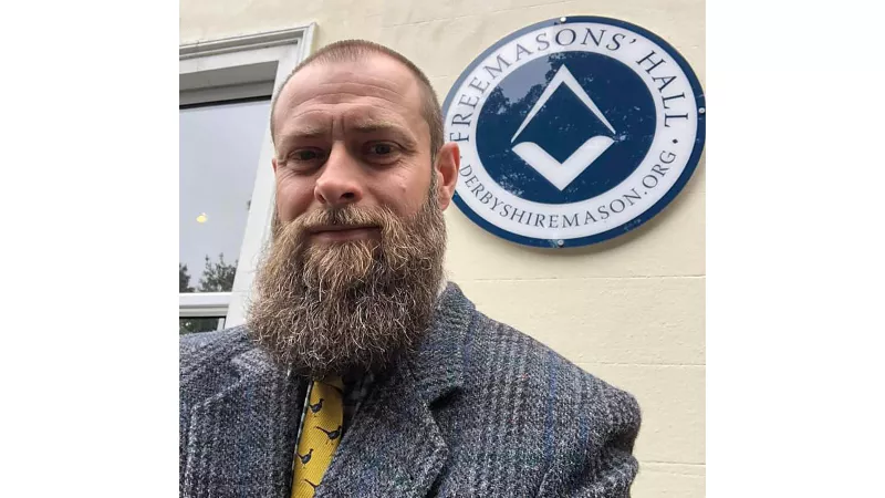Matthew Wainwright is stood in front of the sign for Derbyshire Freemasons' Hall. He has a large beard and is wearing a grey suit.