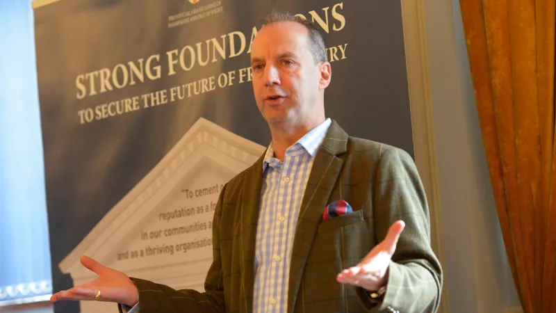 John Whitaker is photographed while giving a speech, he is wearing a green tweed jacket on top of a blue gingham shirt.