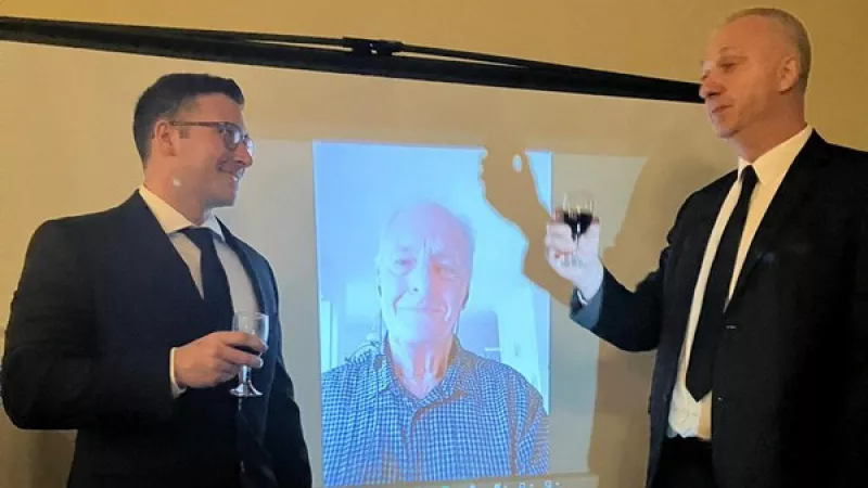 three men toasting, one is participating via video call