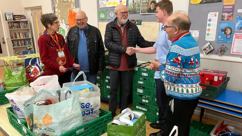 A woman and 5 men standing behind a table which has crates and banks of food items that have been donated to the food bank