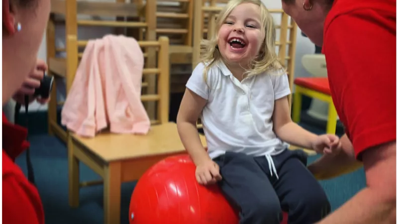 Grace, a young blonde girl, sits on a red yoga ball, she is laughing.