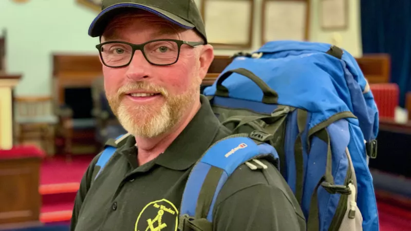 A bearded man with classes wearing a green cap and polo shirt smiling at the camera. He is wearing a blue backpack.