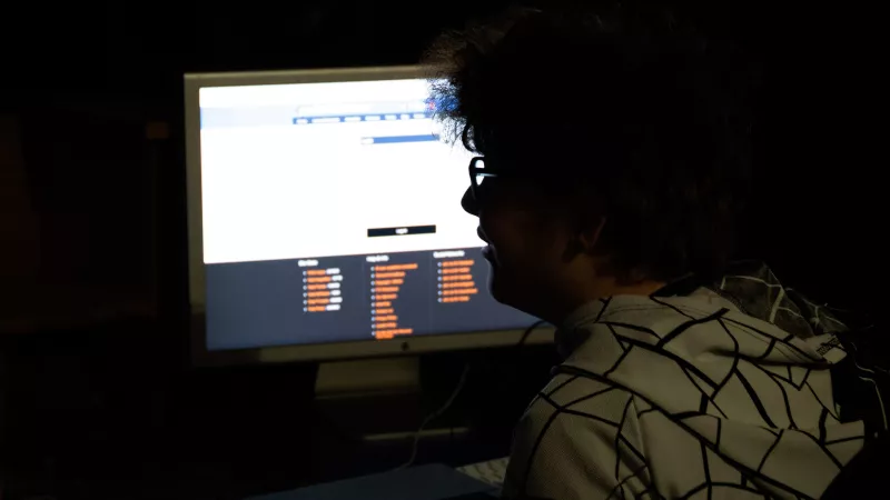 A young man with dark curly hair, sits in front of a computer screen. The light of the screen is bright and leaves the young man in shadow.
