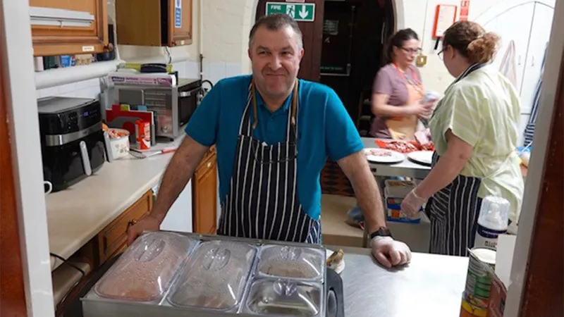 Man stood in kitchen area with covered catering dishes of food in front of him.